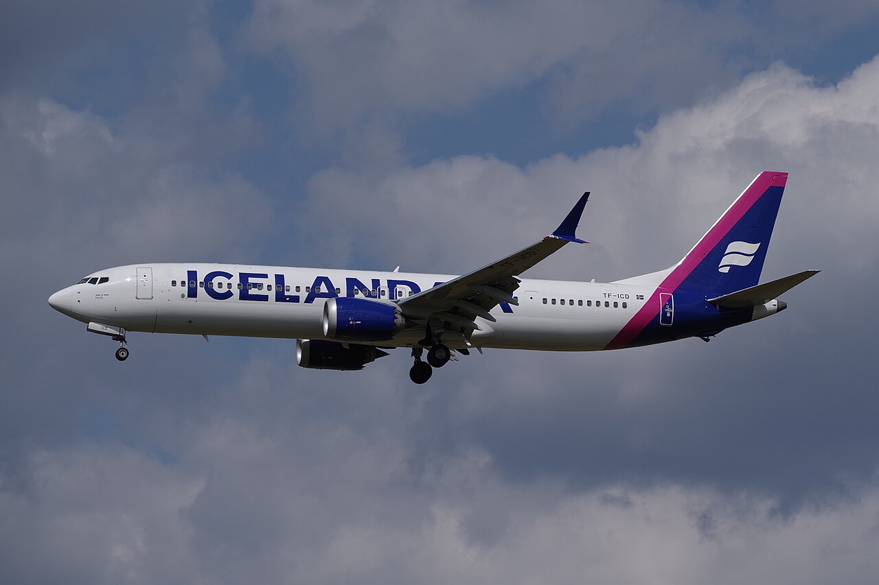 Icelandair B737-9 flying under a cloudy sky, but with some blue peeking through the clouds. The livery is mostly white, but it has pink and dark blue accents.
