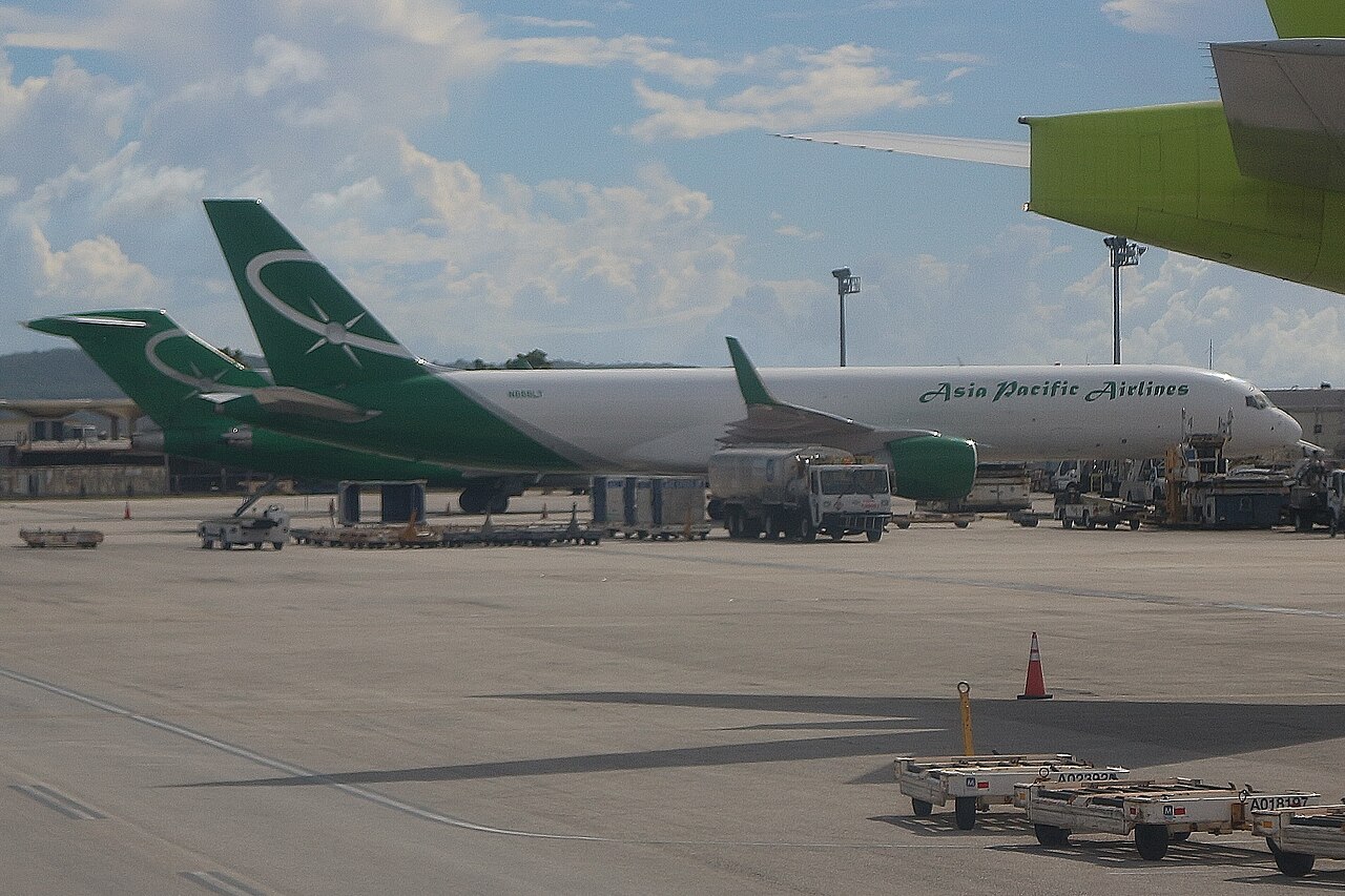 Asia Pacific Airlines cargo aircraft parked on an airport apron with ground service vehicles and cargo equipment nearby under a partly cloudy sky.