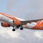 An orange-and-white easyJet passenger airplane taking off into a cloudy sky with its landing gear still extended.