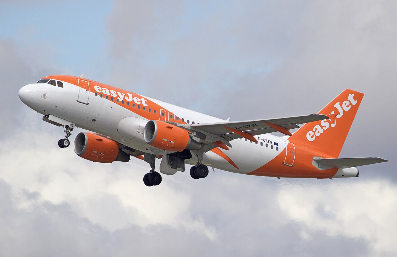An orange-and-white easyJet passenger airplane taking off into a cloudy sky with its landing gear still extended.