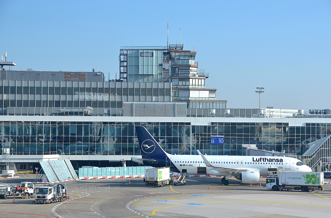 Lufthansa Airbus aircraft parked at an airport gate during ground handling operations, with service vehicles and terminal building in the background.