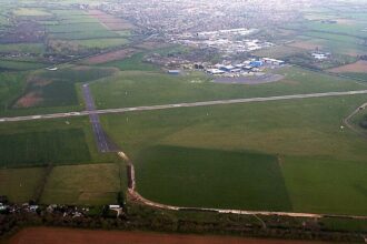 View of a runway and surrounding airfield buildings at London Oxford Airport under a clear sky.
