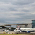British Airways passenger aircraft parked and taxiing near Heathrow Airport terminal buildings, with a plane taking off in the background beneath cloudy skies.