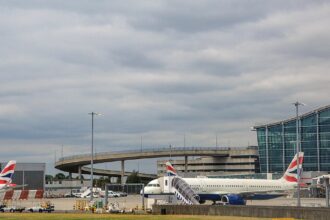 British Airways passenger aircraft parked and taxiing near Heathrow Airport terminal buildings, with a plane taking off in the background beneath cloudy skies.
