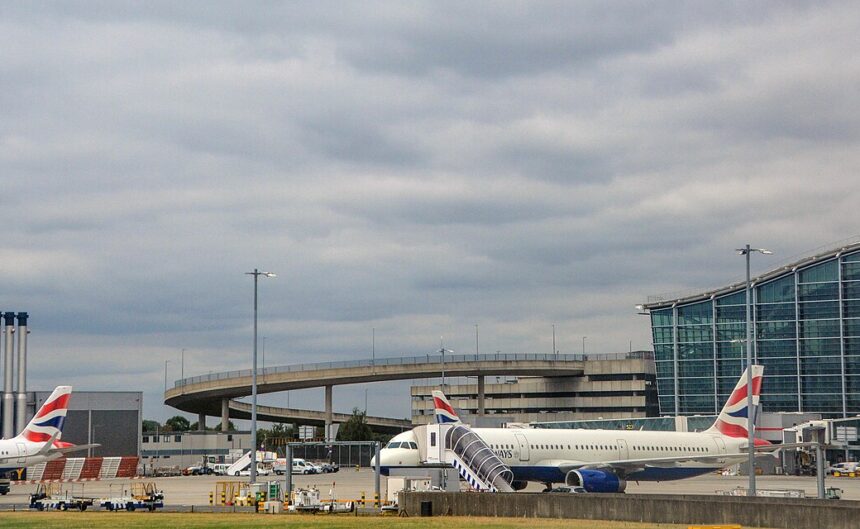 British Airways passenger aircraft parked and taxiing near Heathrow Airport terminal buildings, with a plane taking off in the background beneath cloudy skies.