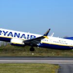 A Ryanair, ultra-low-cost airline, B737 MAX departing Manchester International Airport. The aircraft is under a blue sky, and its livery is white, blue and yellow.