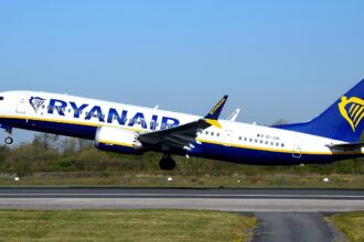A Ryanair, ultra-low-cost airline, B737 MAX departing Manchester International Airport. The aircraft is under a blue sky, and its livery is white, blue and yellow.