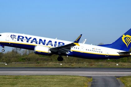 A Ryanair, ultra-low-cost airline, B737 MAX departing Manchester International Airport. The aircraft is under a blue sky, and its livery is white, blue and yellow.