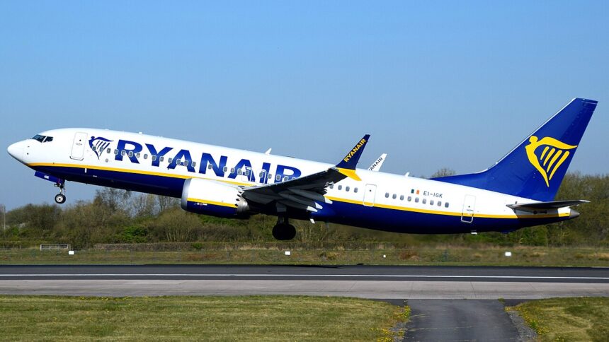 A Ryanair, ultra-low-cost airline, B737 MAX departing Manchester International Airport. The aircraft is under a blue sky, and its livery is white, blue and yellow.