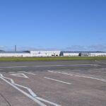 Exterior view of the Spirit AeroSystems manufacturing facility in Belfast, showing a large industrial aerospace plant building beside an airport.