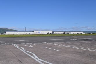 Exterior view of the Spirit AeroSystems manufacturing facility in Belfast, showing a large industrial aerospace plant building beside an airport.