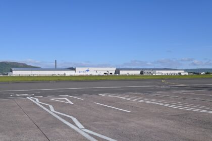 Exterior view of the Spirit AeroSystems manufacturing facility in Belfast, showing a large industrial aerospace plant building beside an airport.