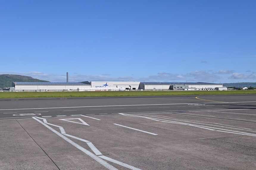Exterior view of the Spirit AeroSystems manufacturing facility in Belfast, showing a large industrial aerospace plant building beside an airport.