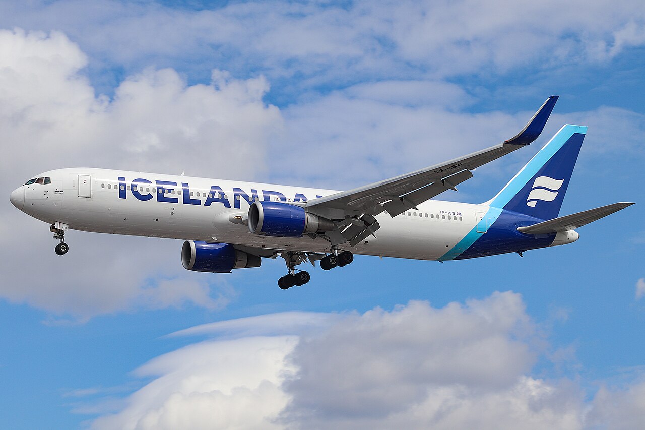 An Icelander B767 flying over London Heathrow under a blue sky with some clouds. The aircraft's outside is white, with its tail being dark and light blue.