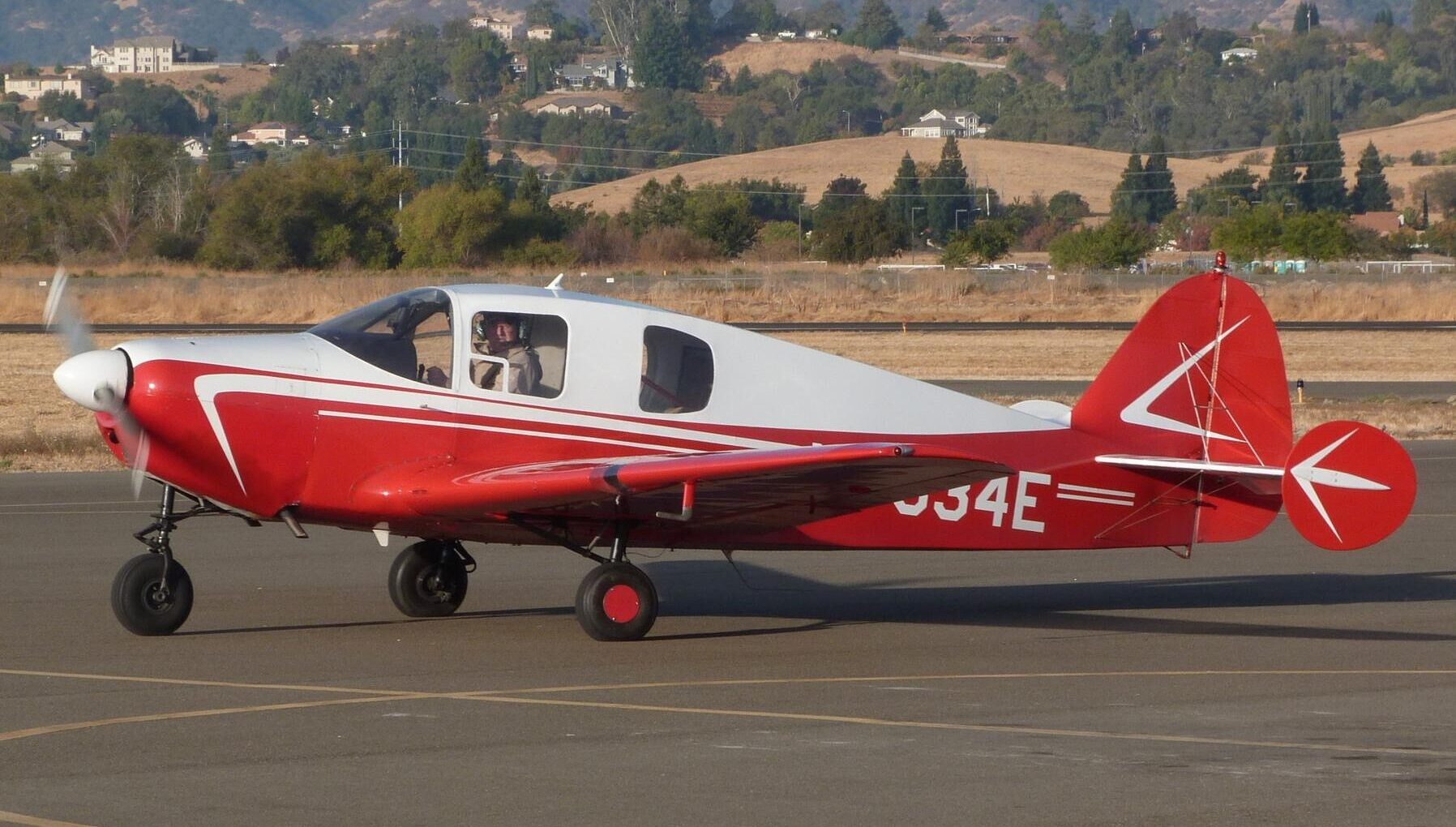A Bellanca 14-19-3 Model 260 on the tarmac. White and red livery, a pilot is seen in the cockpit. Trees, hills and buildings seen in the background.