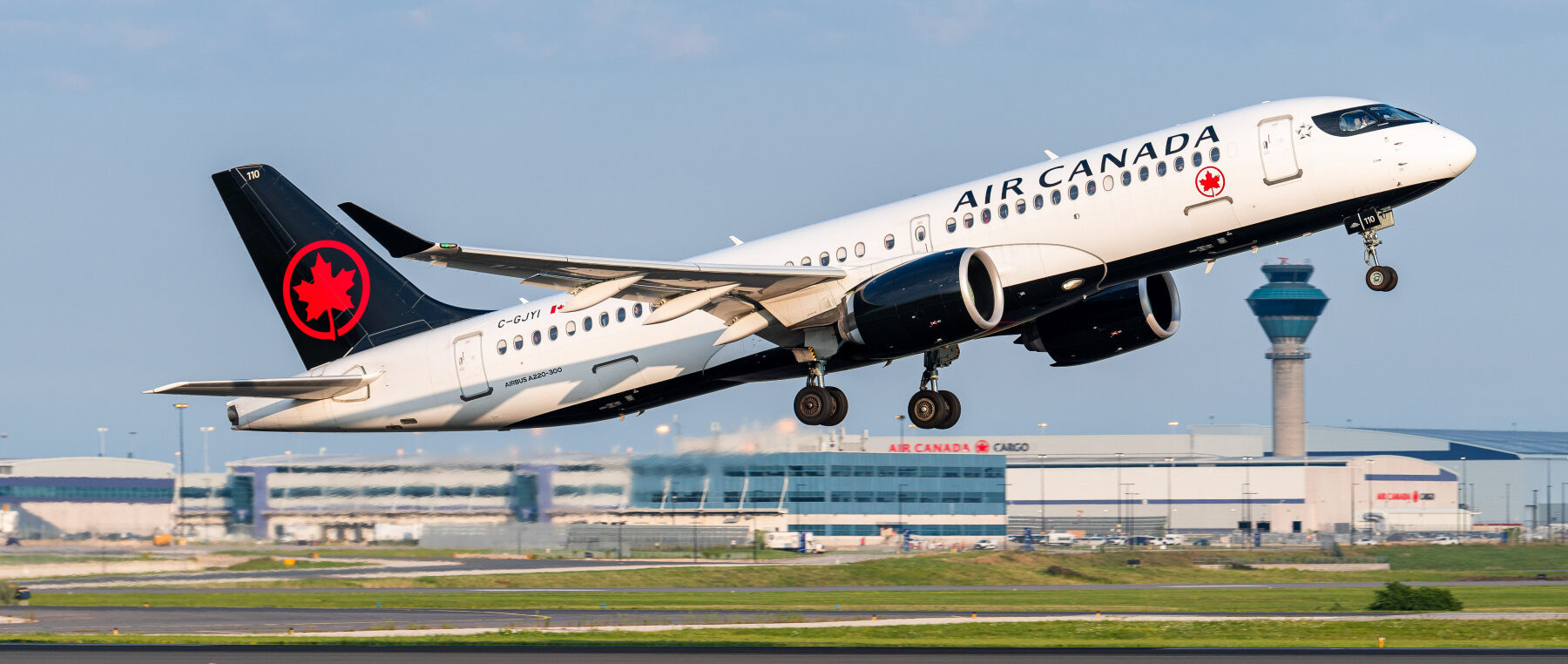 Air Canada Airbus A220 taking off from the runway with buildings and a blue sky in the background.