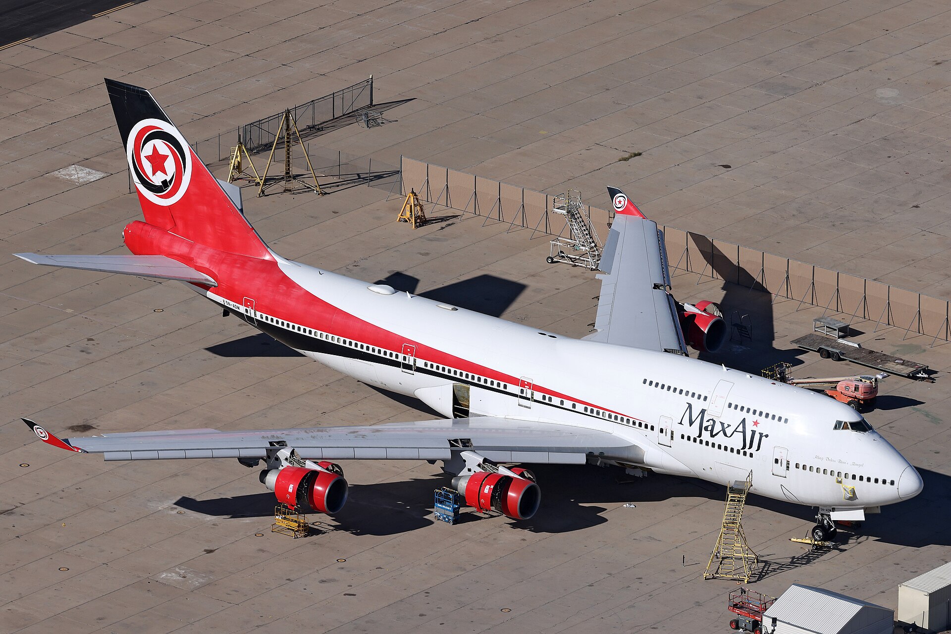 A large double-decker Boeing 747 Max Air plane stands stationary outside an air hangar. The nose is positioned in the bottom right of the image and the tail in the top left. The aircraft is white with the black 'Max Air' branding on the side near the front. The tail of the aircraft is black and red, and has the Max Air logo on it, which is a red star in a black, red, and white swirl. 