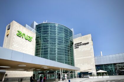 External entrance of Lisbon Airport on a sunny day. The image is taken with a wide lens, with the main entrance building centre left of the image. The central column of the building is all glass windows. Either side, there are thick sections of white brick. In black writing on the right column is the name of the airport. In the foreground is the road leading up to the airport, and in the background is blue sky.