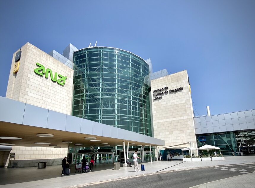 External entrance of Lisbon Airport on a sunny day. The image is taken with a wide lens, with the main entrance building centre left of the image. The central column of the building is all glass windows. Either side, there are thick sections of white brick. In black writing on the right column is the name of the airport. In the foreground is the road leading up to the airport, and in the background is blue sky.