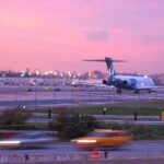 An AirTran B717 aircraft preparing for take off from runway 4 at LaGuardia Airport in New York at sunset. The aircraft is facing away from the camera at a distance, occupying the centre right of the image , travelling away from the camera. The body of the aircraft is white with a blue T shaped tail. Int he foreground is a road with trees and gras either side of it, which is in front of the fence that borders the runway. The sky is pink and purple.