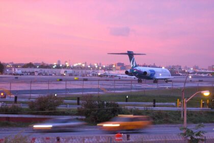 An AirTran B717 aircraft preparing for take off from runway 4 at LaGuardia Airport in New York at sunset. The aircraft is facing away from the camera at a distance, occupying the centre right of the image , travelling away from the camera. The body of the aircraft is white with a blue T shaped tail. Int he foreground is a road with trees and gras either side of it, which is in front of the fence that borders the runway. The sky is pink and purple.