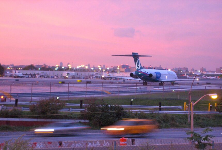An AirTran B717 aircraft preparing for take off from runway 4 at LaGuardia Airport in New York at sunset. The aircraft is facing away from the camera at a distance, occupying the centre right of the image , travelling away from the camera. The body of the aircraft is white with a blue T shaped tail. Int he foreground is a road with trees and gras either side of it, which is in front of the fence that borders the runway. The sky is pink and purple.