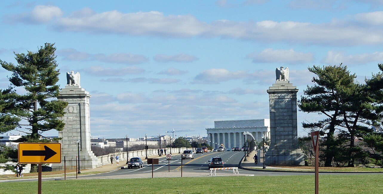 Arlington Memorial Bridge is pictured from a grassy space looking towards the entrance with the Lincoln Memorial in the distance. The entrance to the bridge is adorned with white brick pillars either side with eagle statues atop them. There are trees either side of the pillars. Cars drive onto and off of the tarmac road on the bridge. In the bottom foreground is green grass and in the top background is blue sky. 