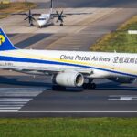 A Boeing 757 occupies the centre of the image, nose facing right and tail facing left. The body of the aircraft is white, with blue branding for Chinese Postal Airlines along the side of the body. The tail is dark blue with yellow logo branding. The aircraft is grounded on a tarmac runway. In the foreground is grass, and in the background is further runways/taxiways