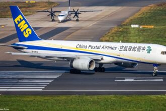 A Boeing 757 occupies the centre of the image, nose facing right and tail facing left. The body of the aircraft is white, with blue branding for Chinese Postal Airlines along the side of the body. The tail is dark blue with yellow logo branding. The aircraft is grounded on a tarmac runway. In the foreground is grass, and in the background is further runways/taxiways