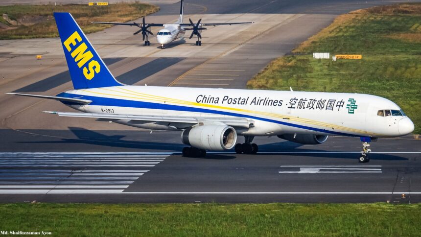A Boeing 757 occupies the centre of the image, nose facing right and tail facing left. The body of the aircraft is white, with blue branding for Chinese Postal Airlines along the side of the body. The tail is dark blue with yellow logo branding. The aircraft is grounded on a tarmac runway. In the foreground is grass, and in the background is further runways/taxiways