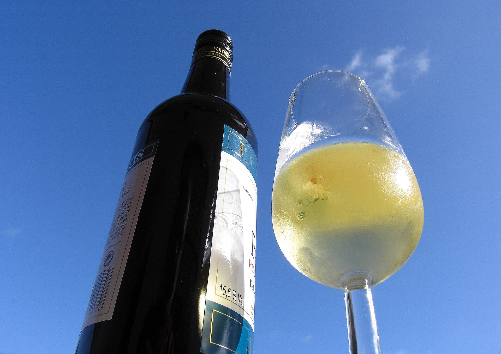 A wine bottle stands centre left next to a wine glass filled halfway with white wine on the centre right. The image is taken from slightly below angled looking up at the wine. the background is a blue sky with minimal clouds, a nice sunny day.