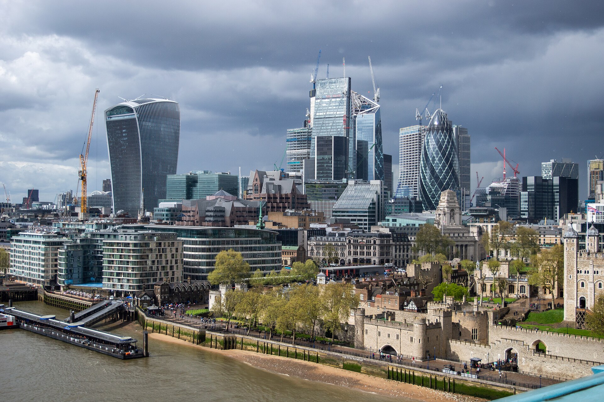 A view of London from London Bride during an overcast day. The Thames can be seen in the lower left corner. Large buildings occupy the centre, including famous landmarks like the Shard, from the left to the right. The background is occupied by moody grey clouds. 