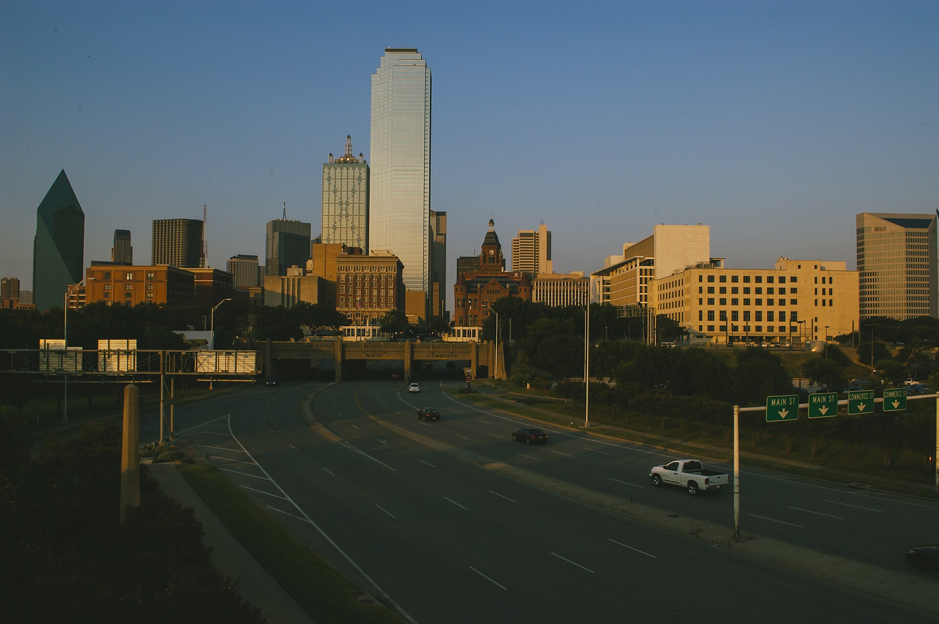 Skyline of Dallas, Texas in the evening at sunset. The bottom half of the image shows a highway in the shade leading up to the city with a few cars dotted around. Along the centre of the image is the city's skyline with a range of buildings of different heights, colours, shapes, and functionalities, all brightened in the sunset. The top section of the image shows a clear blue sky.