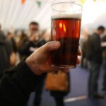 In the upper right centre a pint of beer is positioned, held by a Caucasian hand in the centre leading into an arm dressed in black long sleeve in the left corner. The background is stylistically blurred, but multiple other people can be seen.