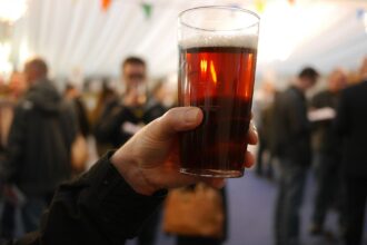 In the upper right centre a pint of beer is positioned, held by a Caucasian hand in the centre leading into an arm dressed in black long sleeve in the left corner. The background is stylistically blurred, but multiple other people can be seen.