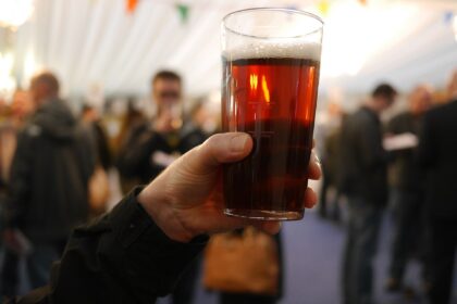 In the upper right centre a pint of beer is positioned, held by a Caucasian hand in the centre leading into an arm dressed in black long sleeve in the left corner. The background is stylistically blurred, but multiple other people can be seen.