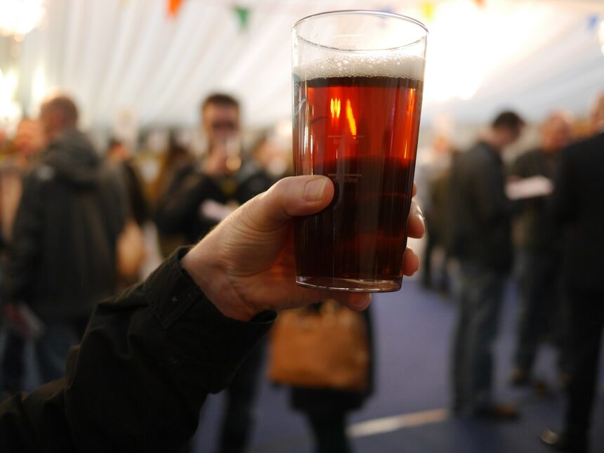 In the upper right centre a pint of beer is positioned, held by a Caucasian hand in the centre leading into an arm dressed in black long sleeve in the left corner. The background is stylistically blurred, but multiple other people can be seen.