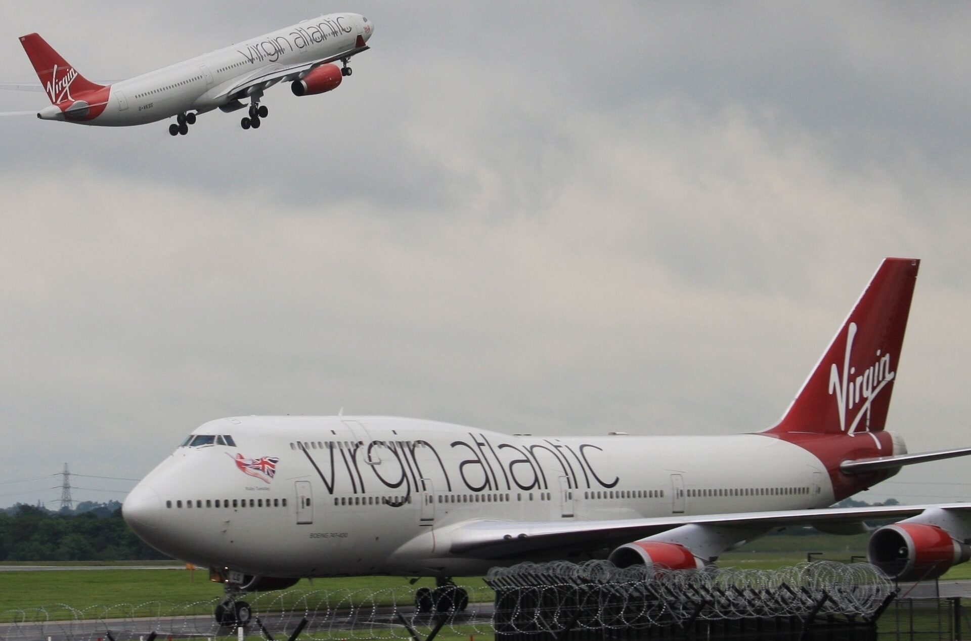 A Virgin Atlantic aircraft is on the runway in the foreground, while another Virgin Atlantic aircraft has taken off in the background, heading up towards the grey-ish sky.