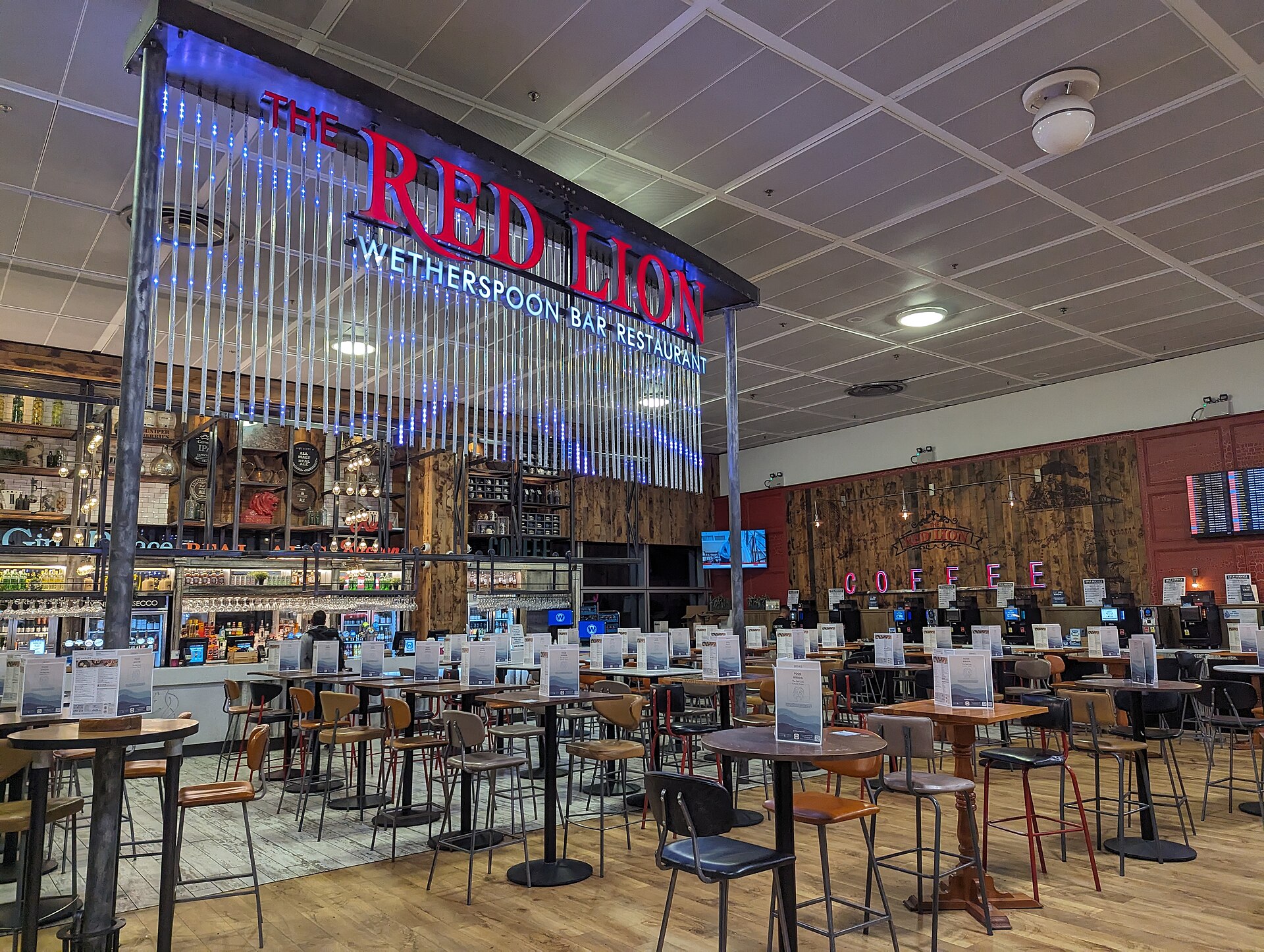 The entrance to the Red Lion Wetherspoons branch in London Gatwick North Terminal. The bar is in the background of the image, along with tables and chairs in brown and black. There also tables and chairs in the foreground. Centre left of the image - the main focus - is the sign that says 'Red Lion' in large red lettering, followed by 'Wetherspoon Bar Restaurant' underneath in smaller white writing, both on a light-up background. This sign is very large, spanning from the ceiling to the middle of the image. 
