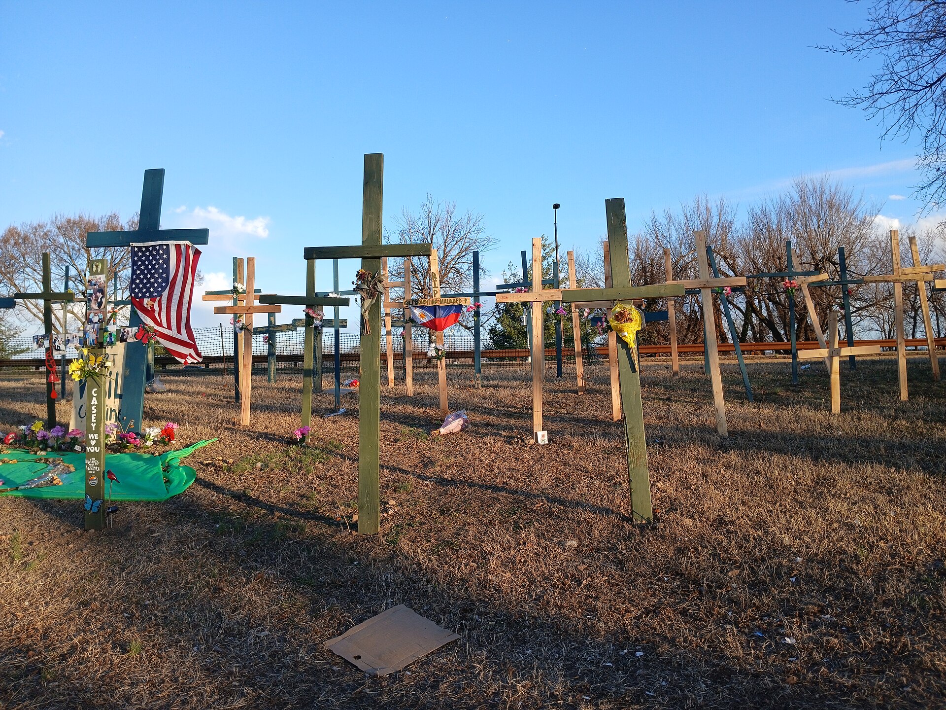 A number of wooden crosses stand in area of brown grass, as a memorial site. The crosses are adorned with American and other nation flags, paint, flowers, messages and more trinkets. In the background are distant trees and blue sky.