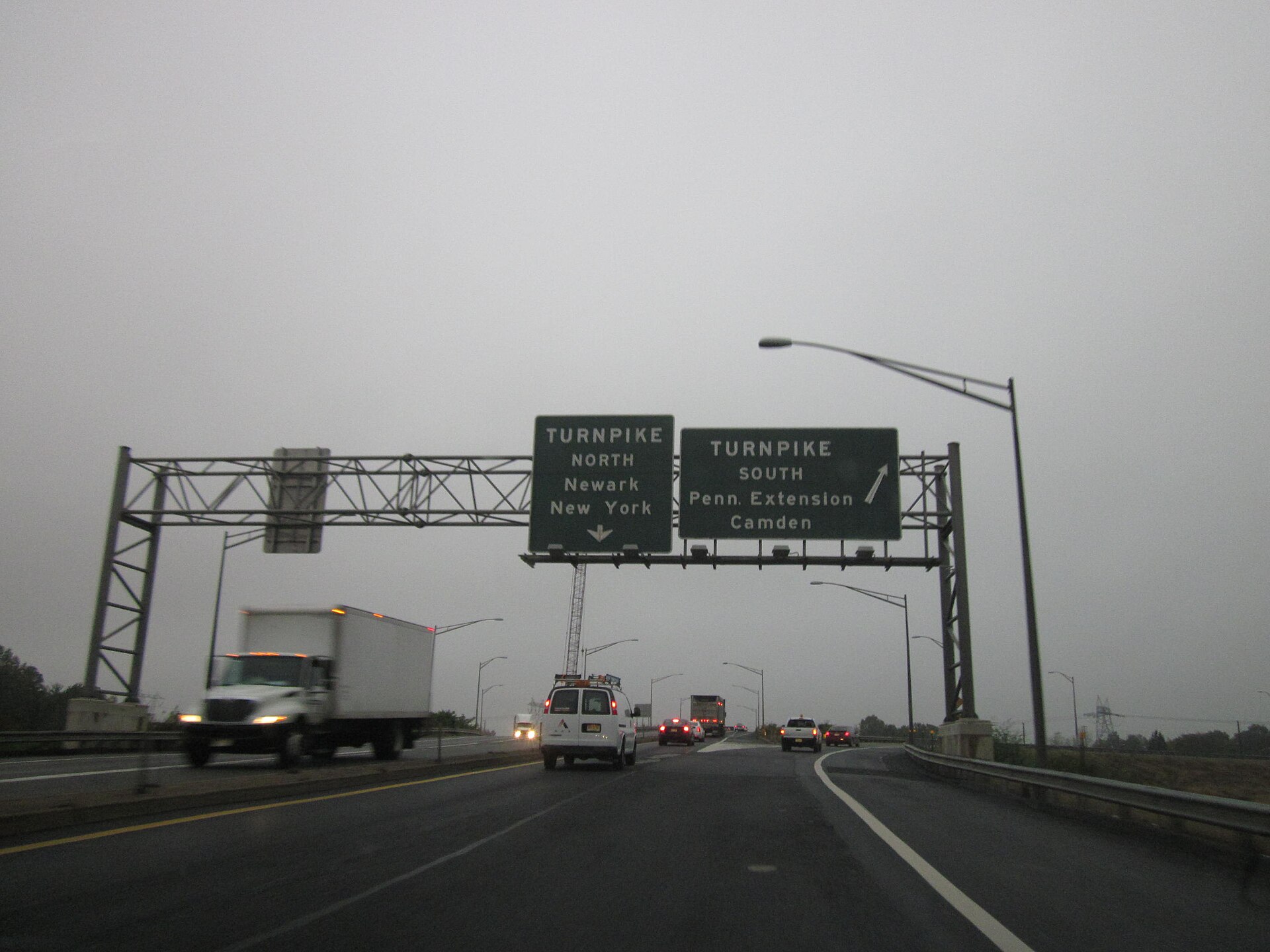 A dark green motorway sign for the New Jersey Turnpike, with blurry moving vehicles on a very grey foggy day. 