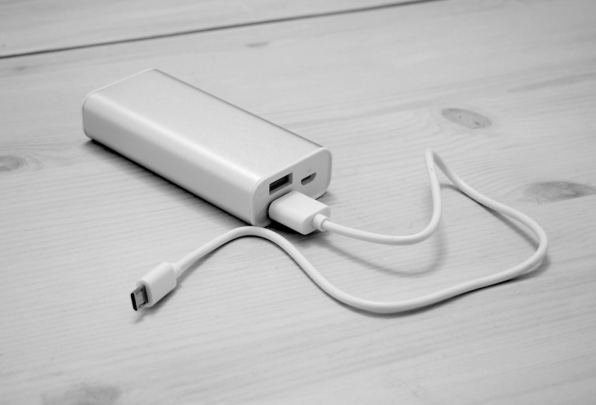 A greyscale black and white image of a power bank on a wooden surface, probably a wooden table. The power bank is white, with three plug in ports, two of which are black because they are empty, and the final one has a white wire plugged in, which protrudes out in a sort of curve. The power bank and wire are in the centre of the image. 