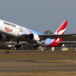 In the centre a Qantas Airline aircraft ascends in take off, nose facing upper left. The bottom of the image shows a tarmac runway and yellow grass. The background is a blue-grey sky. The aircraft is a Disney edition plane, with characters printed on the side and back end of the plane, with the baby blue and red branding. The aircraft is a Boeing 767-300
