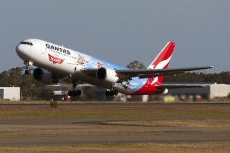 In the centre a Qantas Airline aircraft ascends in take off, nose facing upper left. The bottom of the image shows a tarmac runway and yellow grass. The background is a blue-grey sky. The aircraft is a Disney edition plane, with characters printed on the side and back end of the plane, with the baby blue and red branding. The aircraft is a Boeing 767-300
