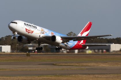 In the centre a Qantas Airline aircraft ascends in take off, nose facing upper left. The bottom of the image shows a tarmac runway and yellow grass. The background is a blue-grey sky. The aircraft is a Disney edition plane, with characters printed on the side and back end of the plane, with the baby blue and red branding. The aircraft is a Boeing 767-300