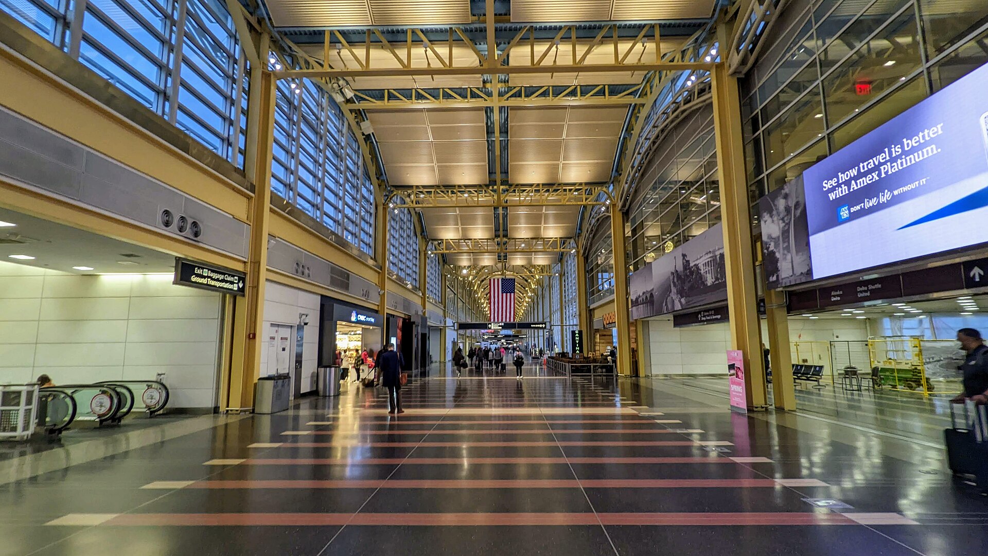 The inside of a Ronald Reagan Washington National Airport (DCA) terminal. The picture is taken in the middle of a walkway looking down the path. Yellow beams arch the walkway, with a help desk and large information screen to the right of the image and escalators down before some shops to the left. In the centre hanging down from one of the arches is an American flag. People walk and wait with luggage. 