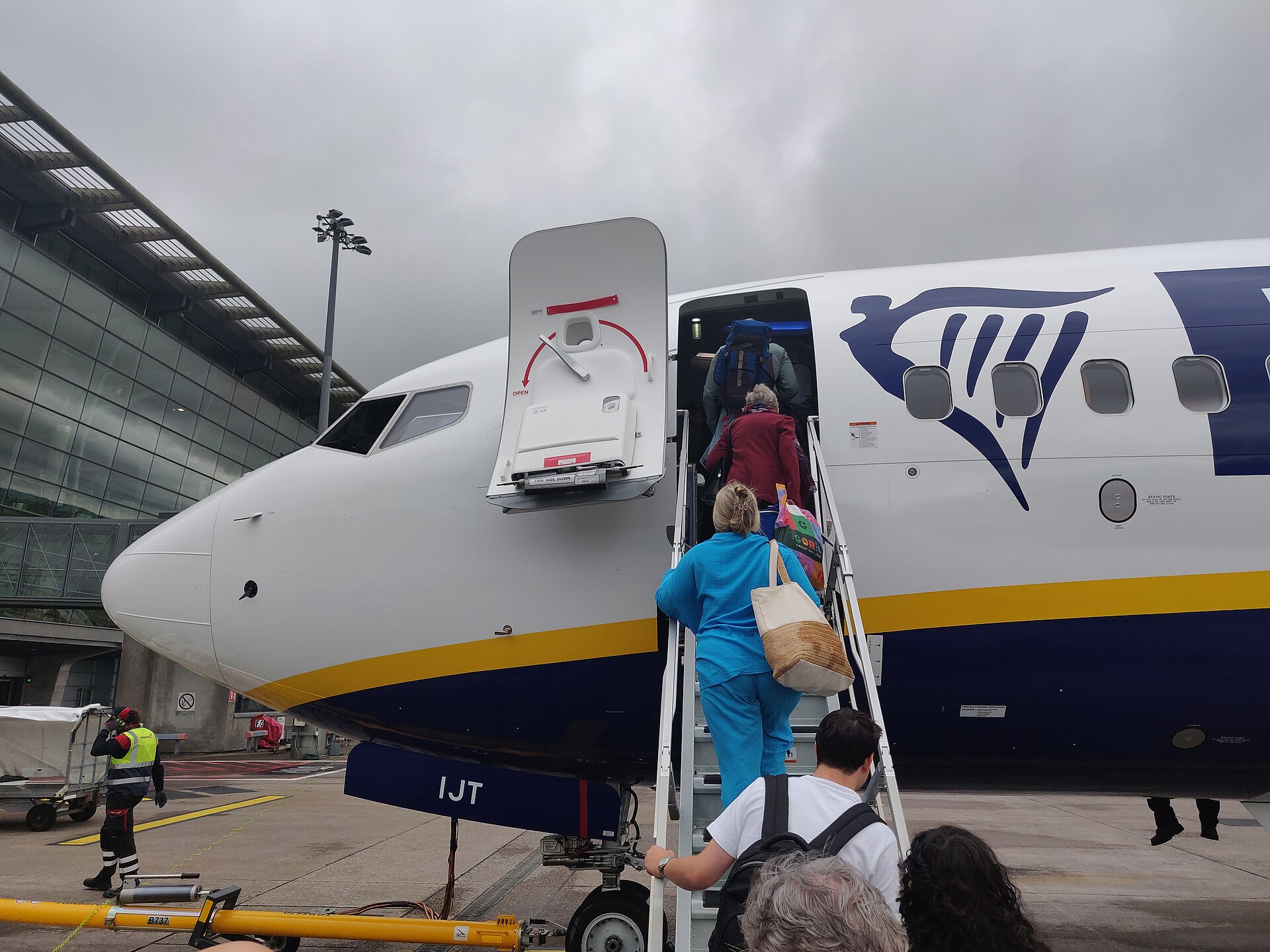 Positioned across entire centre is the front of a Ryanair plane, the nose facing to the left. The door is open with stairs leading up to it, and a queue of people board the flight carrying hand luggage. The aircraft is white with the navy blue and yellow branding across the underbelly, and the dark blue Ryanair logo can be seen to the right of the door. In the background, the sky is overcast grey and cloudy. A terminal building can be seen in the left background, with a member of ground staff below the nose in high visibility gear. 