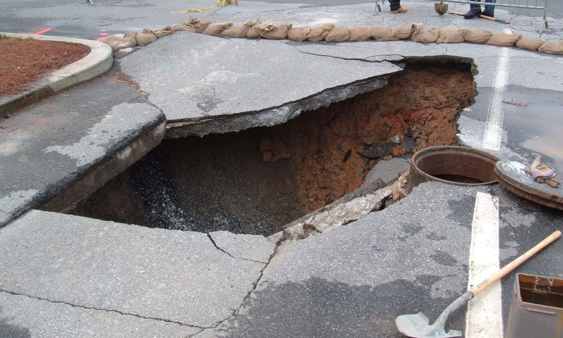 In the centre of the image is a sinkhole in a tarmac road. The inside of the sinkhole is brown and grey and shadowed, and surrounding the sinkhole is pale grey tarmac. A pipe is positioned in the right wall of the sinkhole, with a while painted line before and after it. What look like sandbags can be seen placed around the sinkhole.