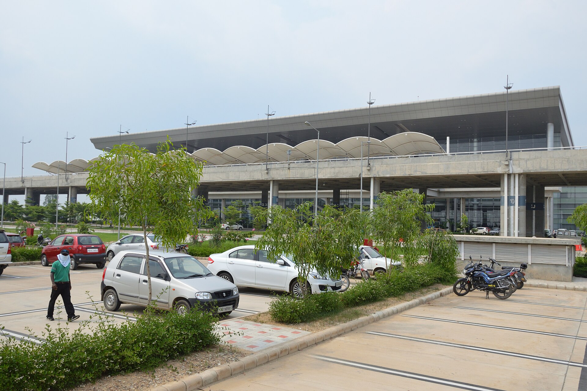 The entrance and car park in front of a terminal at Chandigarh Airport (IXC) from the outside. The building stretches across the centre of the image, with the car park (with several cars parked in it) positioned in the foreground. In the background, the sky is slightly overcast. Bushes and small trees line the boundaries of the car park. 