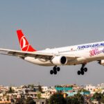 A white Turkish Airlines aircraft is positioned to the centre right of the image, shown in flight. The nose of the aircraft is close to the right edge of the image, the tail close of the centre. The body of the aircraft has Turkish Airlines branding along the side towards the front. The tail of the aircraft is red. The background shows a blue sky and rooftops of buildings.